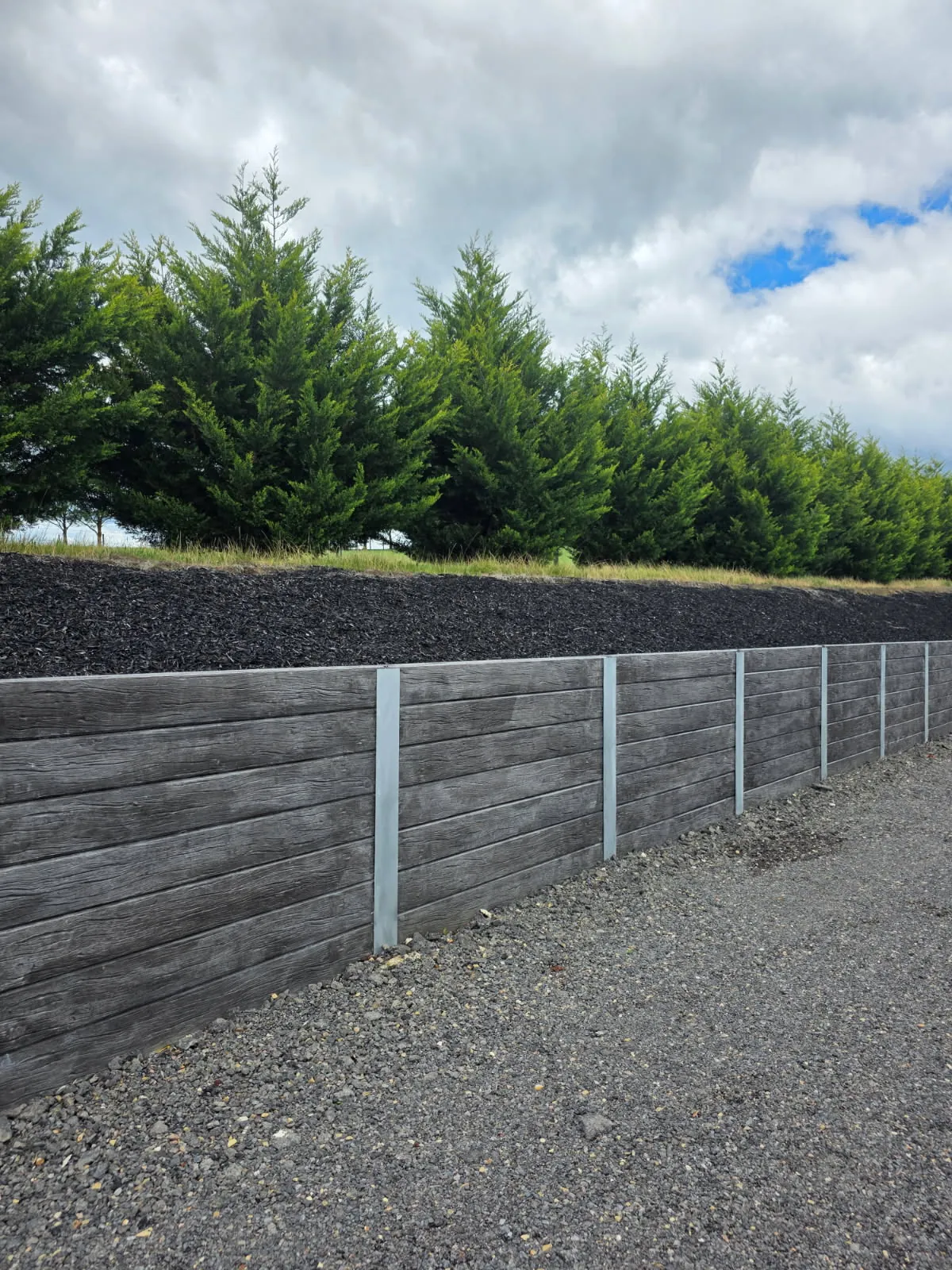 Finished charcoal woodgrain sleeper retaining wall running along a gravel driveway